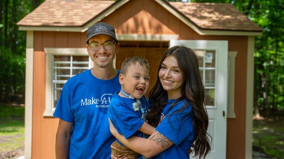 Axle is being held by his parents in front of his new playhouse. They are all smiling and laughing