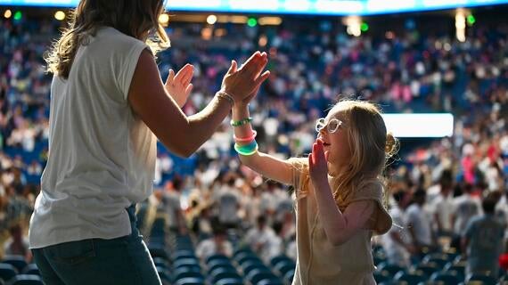 A woman and a young girl high-fiving in an outdoor stadium setting, likely at a concert or event. The girl wears clear-framed glasses, and glow-in-the-dark bracelets on her arm. The background is blurred with people sitting in seats.