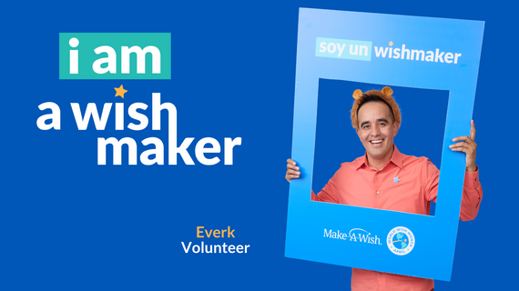 A smiling man with fuzzy ears holds a blue "soy un wishmaker" photo booth frame, standing in front of a blue background with the text "i am a wish maker" and "Everk Volunteer" on the left.
