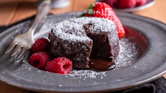 Chocolate lava cake sprinkled with powdered sugar on a dark plate with raspberries, a strawberry, and a fork