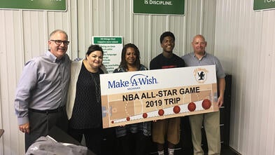 Four adults stand with a teenage wish kid in front of a wall that has many signs with inspirational sayings. The group of people holds a large countdown sign with the Make-A-Wish Michigan and All Weather Seal company logos and images of basketballs and a basketball court.