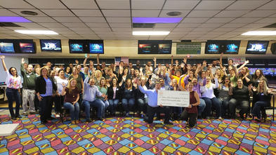 A large group of people gather together in a bowling alley for a photo. Many of them have thrown their hands in the air. In the front of the group, two adults are kneeling and holding an oversize check, made out for sixty thousand dollars to Make-A-Wish Michigan from Arby's.