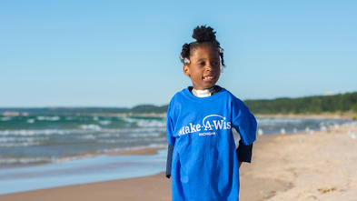 A child with dark curly hair stands on the beach next to the water. The child is wearing a royal blue Make-A-Wish t-shirt and black floral patterned leggings.