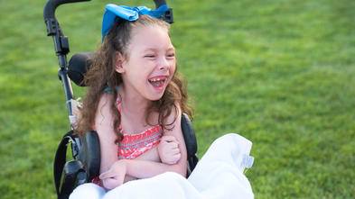 Sitting in a wheelchair with a white blanket across the lap and a big blue bow in curly hair, a 6-year-old child laughs with unrestrained joy. Behind the child is a field of smooth green grass.