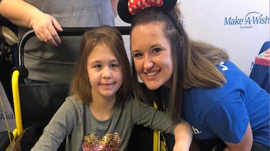 An adult with shoulder-length straight dark hair crouches next to a child sitting in a yellow wheelchair. The adult is wearing a royal blue Make-A-Wish t-shirt, jeans, and Minnie Mouse ears. The child is wearing a long-sleeved gray t-shirt with a multi-colored heart, jeans, and purple cowboy boots and clutching a large brown and white stuffed horse.