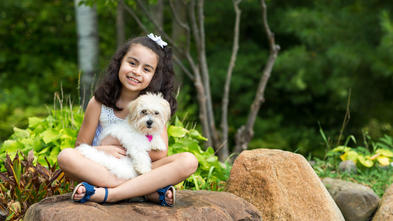 A 7-year-old child with shoulder-length curly dark hair cuddles a fluffy white puppy while sitting cross-legged on a large rock. The child is wearing a white lace tank top and shorts with blue sandals. In the background there are large green plants and trees.