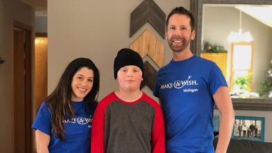 Two adults in royal blue Make-A-Wish t-shirts and jeans stand on either side of a child in a red and gray shirt and black beanie. The background is a living room with a gray couch and wooden wall art.