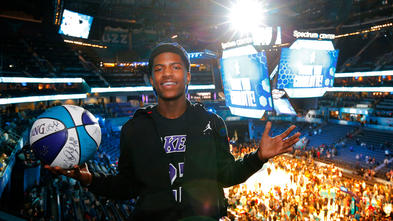 A 17-year-old teenager in a black Lakers t-shirt, partly unzipped black jacket, and backwards baseball cap stands at the top of a basketball court filled with people. In the left hand hand is a signed basketball.