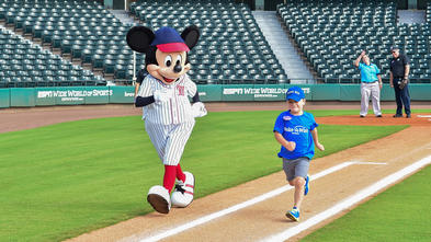A 6-year-old child wearing a blue Make-A-Wish t-shirt and gray shorts runs towards first base on in an empty baseball stadium. Mickey Mouse, who is wearing a blue and white striped baseball uniform and red and blue baseball cap, runs beside the child.