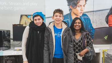 A teenagee with short curly brown hair, wearing a royal blue Make-A-Wish t-shirt, gray winter jacket and black athletic pants, smiles broadly. The teenager stands between two wish-granting volunteers in Best Buy's computer department. On the left, volunteer Timerra is wearing a royal blue Make-A-Wish beanie and a long black scarf. On the right, volunteer Millie is wearing a black and white patterned tunic and holding the receipt from the purchases made at Best Buy. 