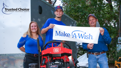 Mason with mom and company rep holding Make-A-Wish sign standing on red lawnmower.