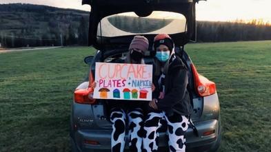 Two masked teens hold a sign offering cupcakes, perched on the tailgate of a car.