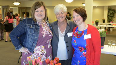 Three adults in bright clothing stand with their arms around each other. In front of them is a large bouquet of red tulips.