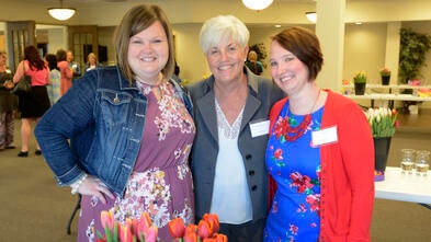 Three adults in bright clothing stand with their arms around each other. In front of them is a large bouquet of red tulips.
