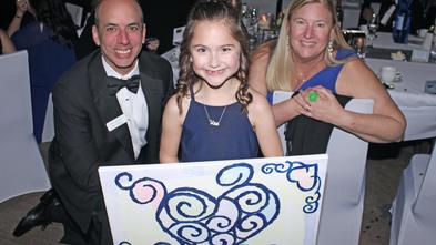 Wish Kid Olivia smiling as she holds her artwork with donors at the Wish Ball