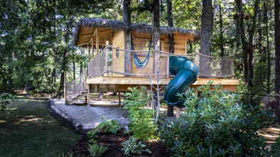 Libby's wooden treehouse nestled among the trees, with stairs and a thatched roof and a green twisty slide