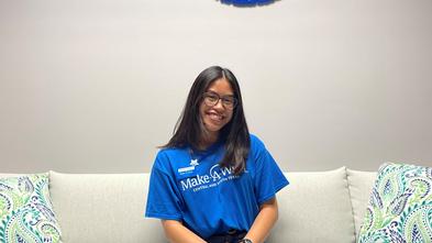 Intern Irissa with a Make-A-Wish shirt on sitting on a couch under a Make-A-Wish sign