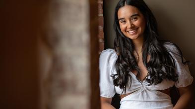 Alexa wears a white shirt with long brown hair and stands against a stone backdrop, smiling at the camera.