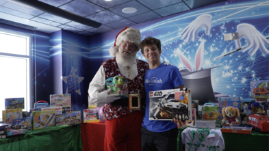 Wish kid in blue shirt standing with Santa in front of toys for Christmas