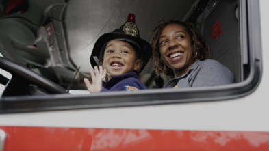 Wish kid in fireman coat and hat with mom waving inside firetruck
