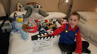 A young boy dressed as Mario sitting on his hotel bed with all his new stuffed animals and toys from the day.