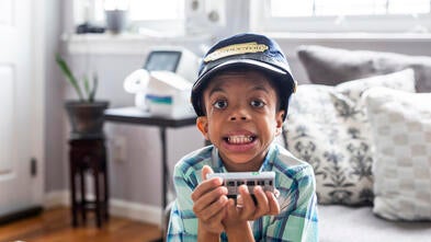 Wish kid Hazael sitting in living room holding toy train with conductor's hat on head