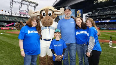 group in Mariners shirt with Mo the Moose mascot on baseball filed looking at camera smiling 