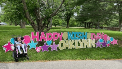 Oaklynn smiling next to a sign celebrating her wish day