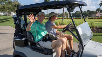 Men on a golf cart waving and smiling on the course.