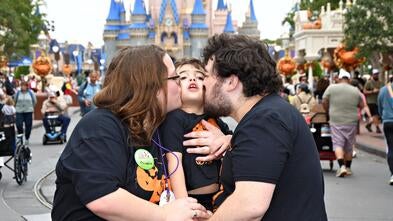 A young boy at Disney with his parents. Both parents are kissing him on the cheek.