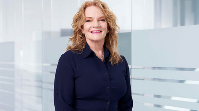 A smiling woman with red curly hair wearing a navy blue collared shirt, posing in front of a frosted glass wall.