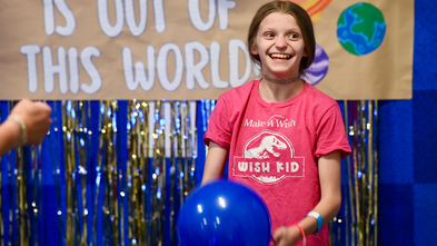 A wish kid stands smiling, holding a blue balloon. They are wearing a pink "Make-A-Wish" shirt with "Wish Kid" printed on it. The background features a banner with "Out of This World" and images of planets, adorned with shiny, gold streamers.