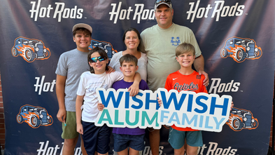 A smiling family of six stands in front of a backdrop featuring the logo "Hot Rods" with car illustrations. Two individuals in the front hold a sign that reads "Wish Alumni Family." 