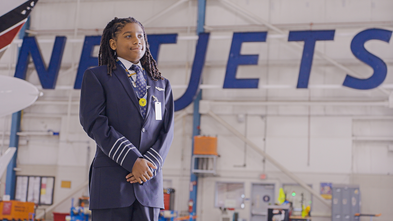 A young Black boy in a pilot uniform stands with folded arms in a large hangar, smiling slightly, with the word "NETJETS" in large blue letters visible in the background.