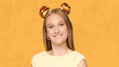 A young woman wearing tiger ears headband smiles as she looks forward against an orange textured background.