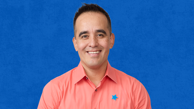 A man with short dark hair and a tan complexion smiles confidently at the camera. He is wearing a coral-colored collared shirt with a small blue star-shaped pin on the left side of his chest. The background is a textured royal blue.