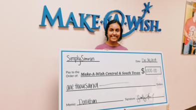 Simran holding a big check made out to Make-A-Wish Central & South Texas for $1,000.