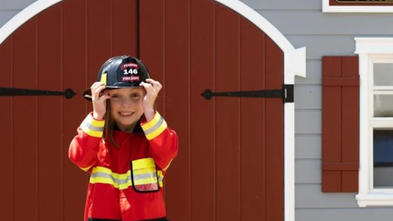 Wish kid Makinley in her firefighting costume standing in front of her firehouse playhouse with a big smile on her face.