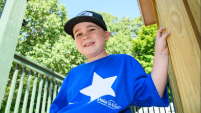 Harrison sits on the porch of his treehouse, smiling, and wearing a bright blue Make-A-Wish t-shirt