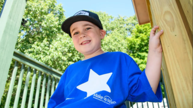 Harrison sits on the porch of his treehouse, smiling, and wearing a bright blue Make-A-Wish t-shirt