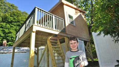 Harrison stands smiling in front of his treehouse, which is brown and has windows and a front porch and a wooden ladder.