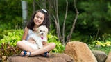 A 7-year-old child with shoulder-length curly dark hair cuddles a fluffy white puppy while sitting cross-legged on a large rock. The child is wearing a white lace tank top and shorts with blue sandals. In the background there are large green plants and trees.