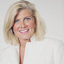 An adult with straight blond hair cut just above their shoulders. The person is wearing a white blazer and clear crystal stud earrings.