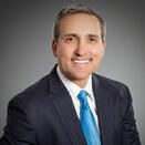 An adult with short gray hair, wearing a black suit jacket with a white shirt and turquoise tie.