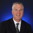 An adult with short gray hair, wearing a black suit jacket with a white shirt and green and navy blue striped tie.