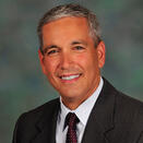 An adult with short gray hair, wearing a gray suit jacket with a white shirt and burgundy tie with small white polka dots.