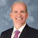 An adult with short gray hair, wearing a black plaid suit jacket with a white shirt and patterned red and royal blue tie.