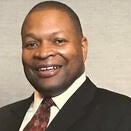Smiling man in corporate headshot photo. Man is wearing a black jacket and white shirt with a red and brown tie.