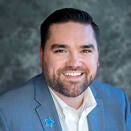 A man smiling. He has dark brown hair, a mustache, and a beard. He is wearing a blue sports jacket with a blue Make-A-Wish star pin on.