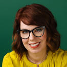 A woman smiling. She is wearing yellow and against a green backdrop. She has dark red hair and is wearing glasses.